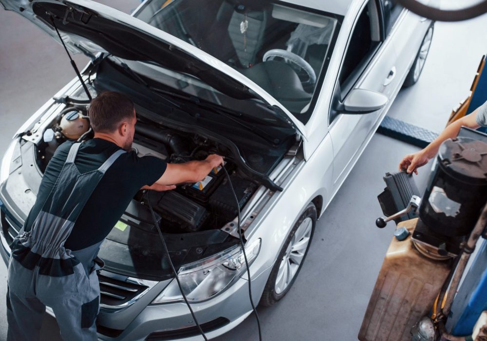 Tests battery. Man in grey uniform repairs white automobile indoors